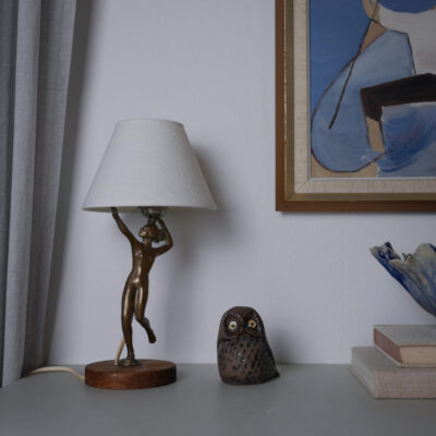 Bronze statue table lamp with a white fabric shade sits on a wooden base, next to a ceramic owl and a small stack of books on a gray surface.