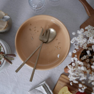 Peach-colored bowl with crumbs and a fork and spoon resting inside, on a dining table viewed from above.