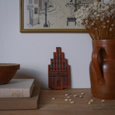 Red terracotta architectural sculpture centered on a wood shelf, with dried flowers in a vase to the right and framed art above.