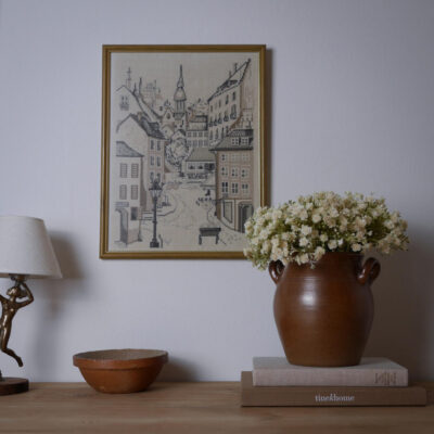 A still-life vignette with a large ceramic vase of white flowers on a stack of books, a decorative lamp, and a framed cityscape hanging on the wall above.