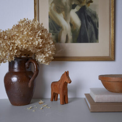 Decorative still life: brown ceramic vase with dried hydrangeas beside a small wooden horse figurine on a table, with framed artwork and bowls in the background.