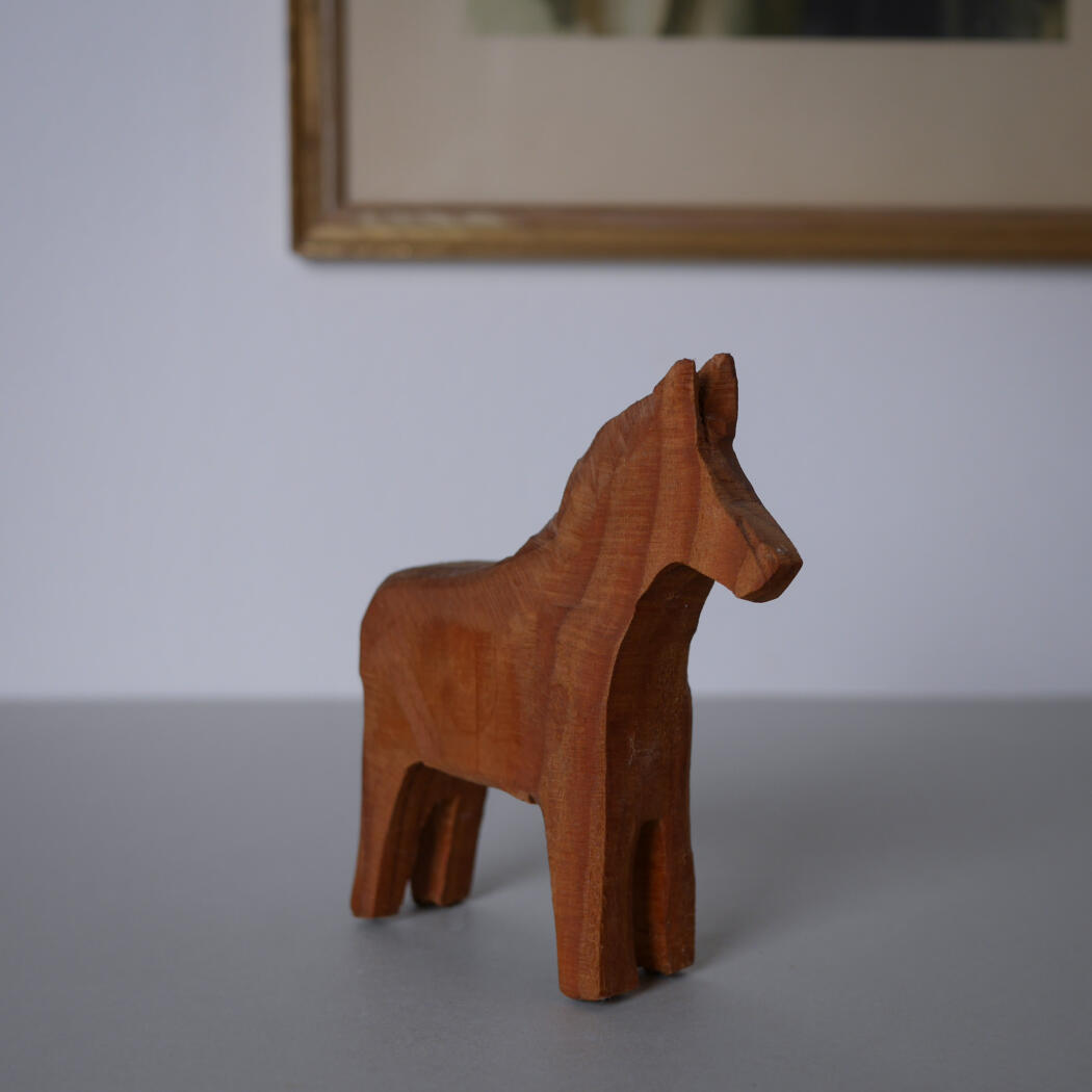Reddish-brown wooden horse carving standing on a gray tabletop with a blurred framed artwork in the background.