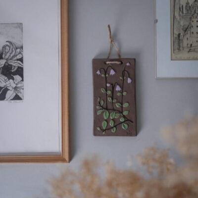 Brown hanging plaque with ceramic purple flowers and green leaves mounted on a wall between framed art pieces