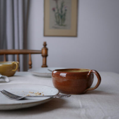 Brown glazed ceramic mug on a white tablecloth with a plate having crumbs and a fork in the foreground; warm, rustic dining room setting in the background.