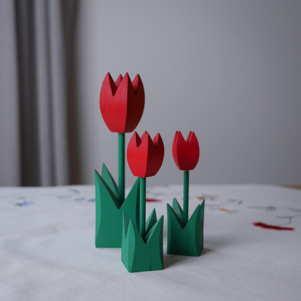 Three red wooden tulips with green stems and leaves on a white tablecloth.