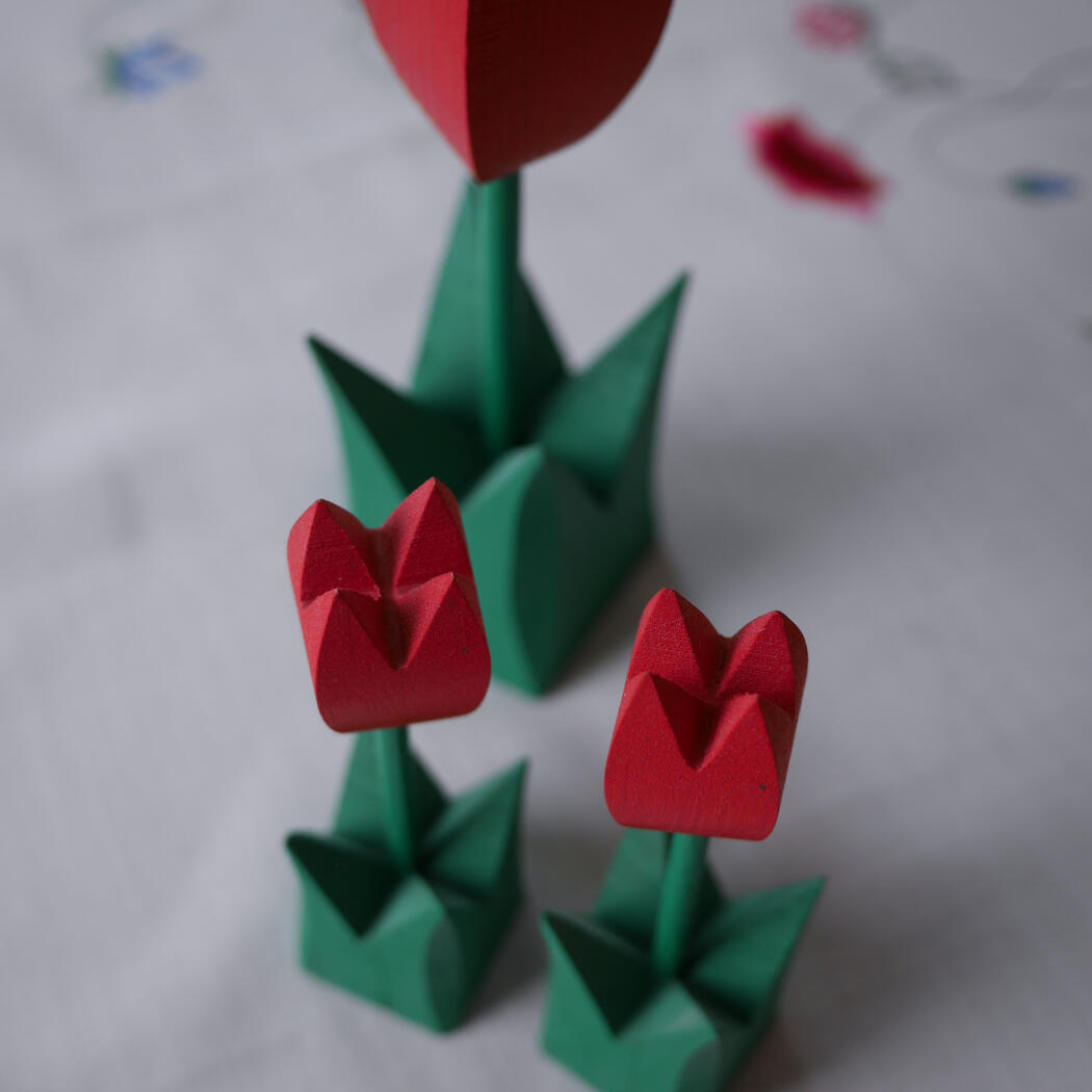 Origami red tulips with green stems and leaves standing in small green bases on a white surface (floral paper art).