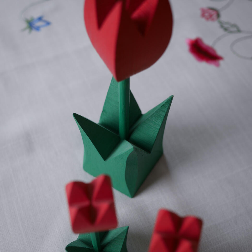 Origami green stems with red folded tulip blooms arranged on a light tablecloth with embroidered pattern in the background.