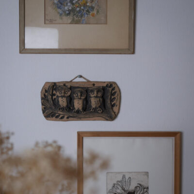 Wall with three decor items: a framed floral print at top, a ceramic plaque with owls mounted in the middle, and a framed botanical drawing at bottom right.