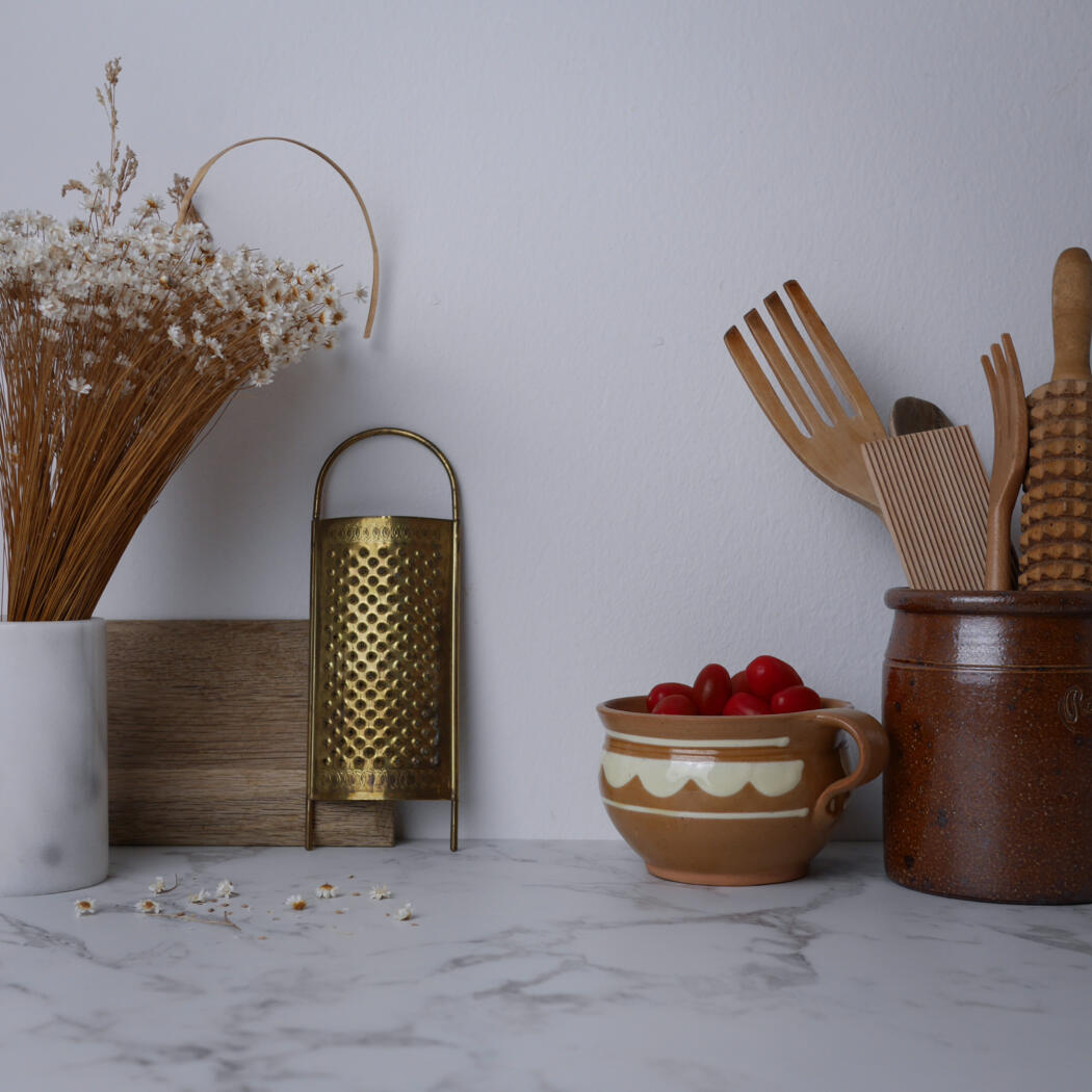 Clay mug filled with red cherry tomatoes sits on a marble countertop beside wooden utensils in a rustic crock and a metal grater; dried flowers in a vase on the left.