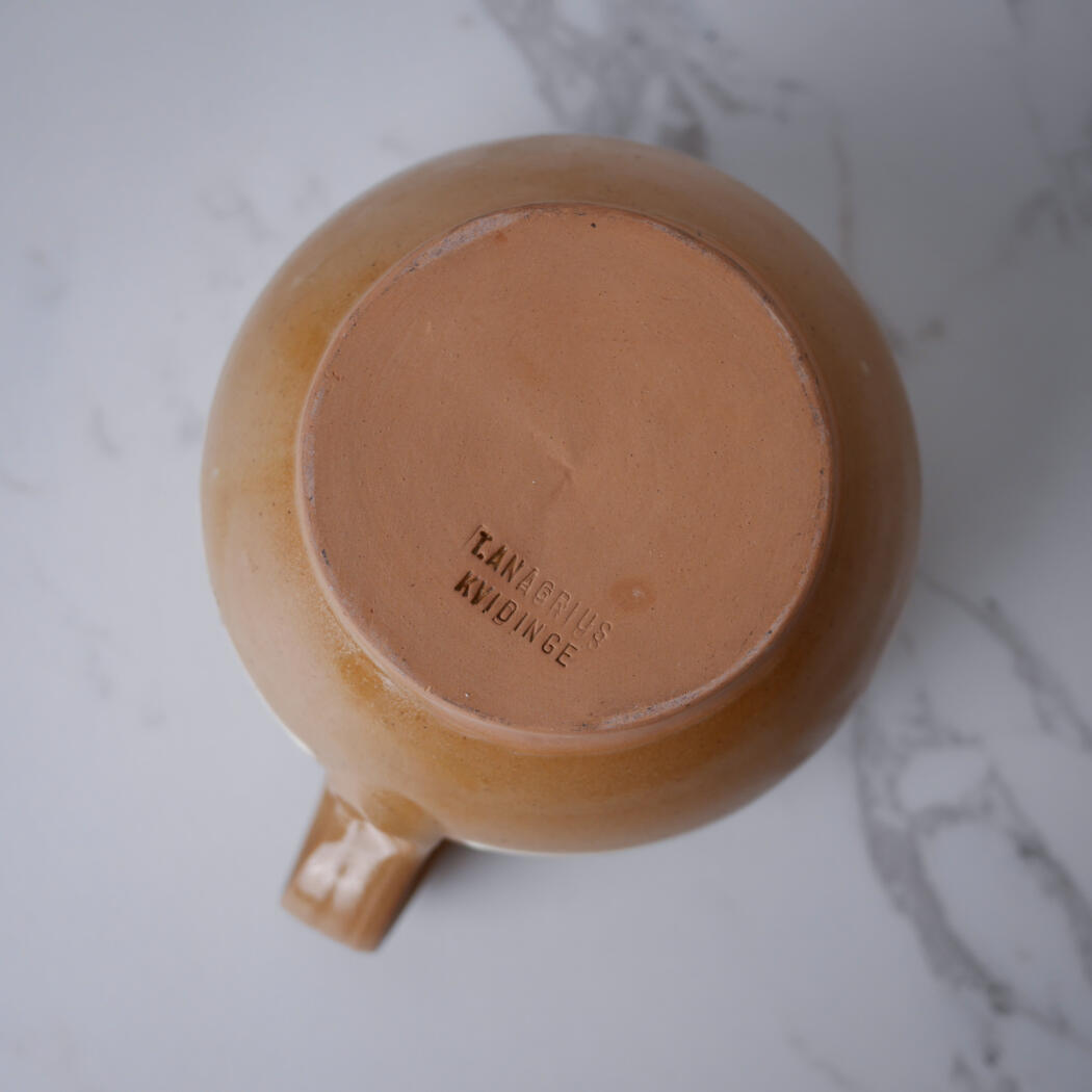 Bottom of a brown ceramic mug showing a maker's stamp on an unglazed base, placed on a light marble surface.