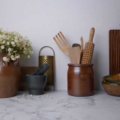 Brown ceramic utensil jar with wooden spatula, fork and brush on a marble counter, beside a mortar and pestle and a brass grater, with white flowers on the left.