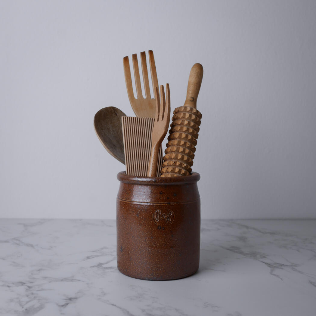 Brown ceramic canister holding wooden kitchen utensils on a marble countertop, against a light gray wall.