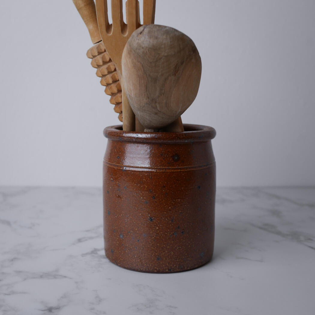 Brown ceramic utensil holder with wooden spoons and spatulas resting inside, placed on a marble countertop against a light gray background
