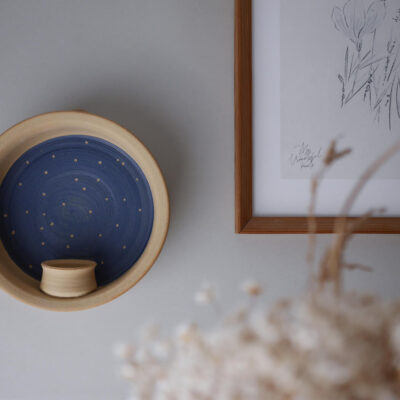 Beige ceramic wall bowl with a blue interior and a small cup inside, hung next to a wooden-framed sketch; dried flowers in the foreground.