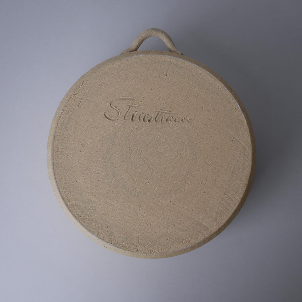 Round beige circular lid or shallow container with a small loop handle at the top, textured surface, and a barely legible cursive inscription embossed on it.