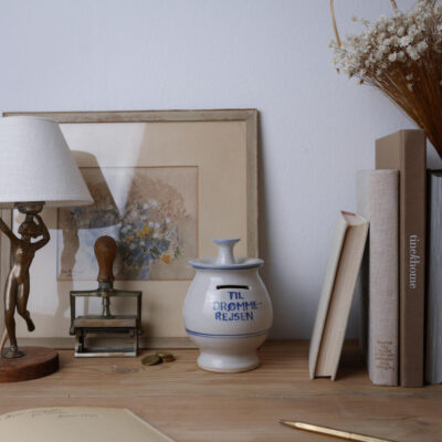 Ceramic jar with blue trim and the Danish inscription 'TIL DROMME-REJSEN' sits on a wooden desk beside neutral books, a vintage lamp, and a dried flower bouquet.