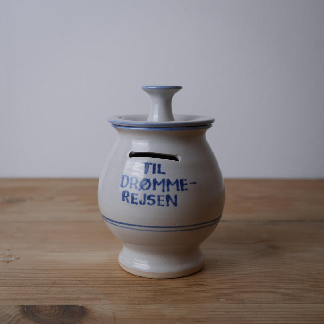 White ceramic jar with a matching lid and blue trim sits on a wooden table; inscription reads 'TIL DRØMME-REJSEN'.
