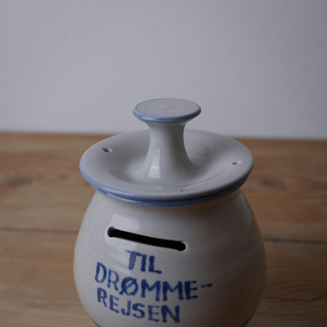 White ceramic savings jar with a coin slot and lid, blue rim, on a wooden table with handwritten blue text on the front.