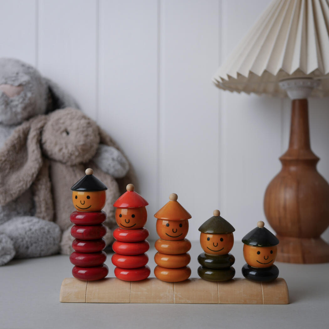 Five colorful wooden stacking dolls with smiling faces and cone hats arranged by height on a light wooden base, background includes a lamp and plush toys in a nursery setting.