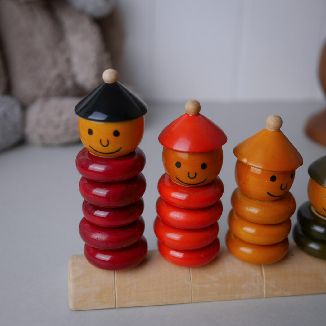Three smiling wooden stacker toys with ring-shaped segments in red and orange, each topped with a colored cone hat, lined up on a light wooden base.