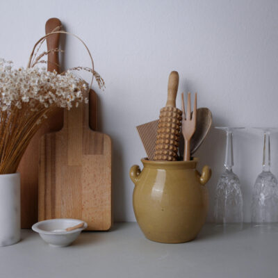 Yellow ceramic jar with wooden utensils beside a wooden cutting board and dried flowers on a white counter.