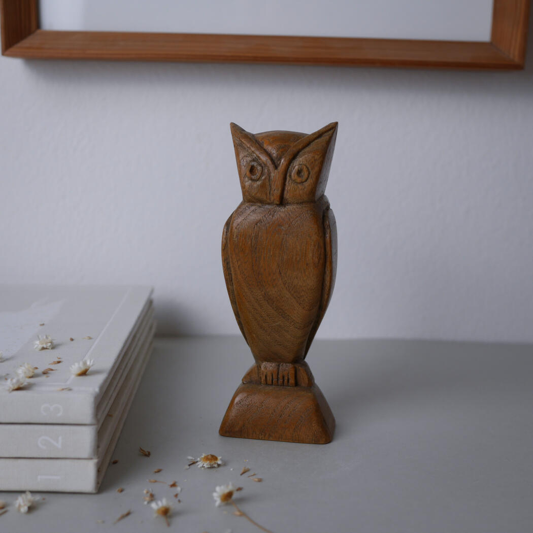 Carved wooden owl statue on a square base, standing on a gray tabletop with a stack of white books to the left and a framed picture above.