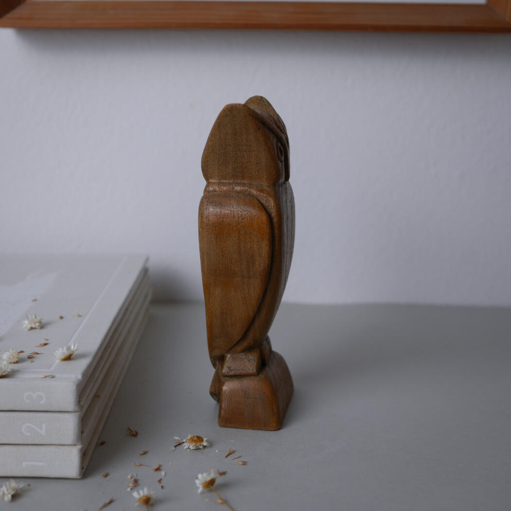 Wooden carved bird statue standing on a small pedestal beside a stack of white notebooks numbered 1–3, with dried daisies scattered on a pale tabletop.