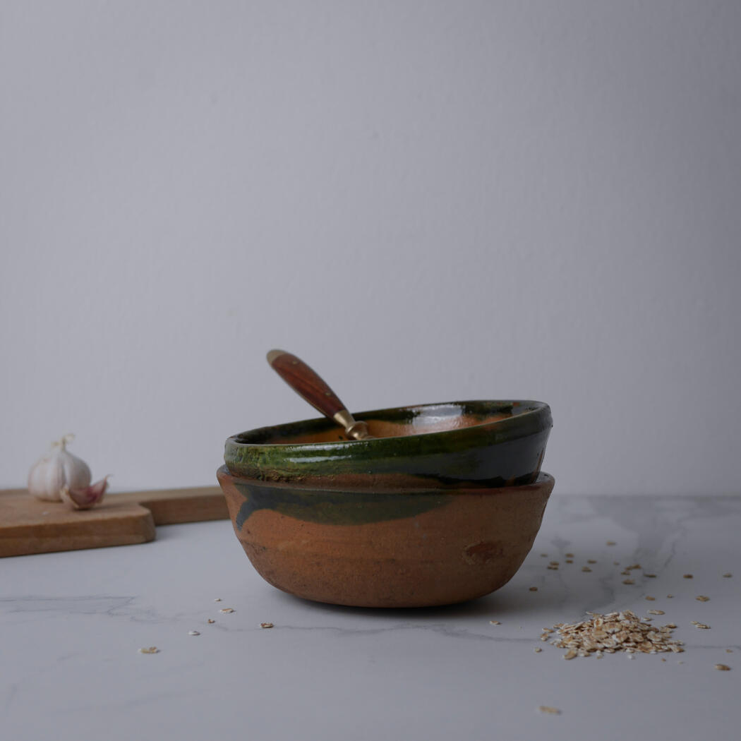 Two stacked rustic ceramic bowls with a wooden spoon resting in the top bowl, on a marble surface with oats spilled nearby and garlic on a cutting board in the background.