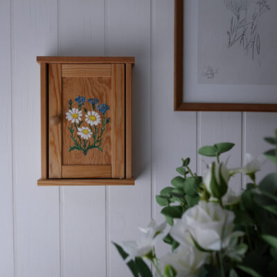 Small wooden wall cabinet with a daisies painting on the door, mounted on a white paneled wall next to a framed sketch; white flowers blur in the foreground.