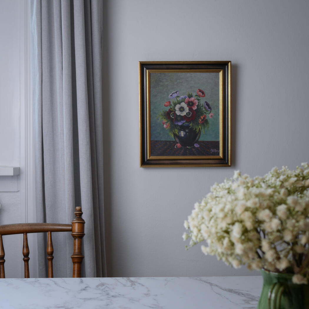 Framed painting of a colorful flower bouquet on a gray wall, with a wooden chair and white marble table foreground.