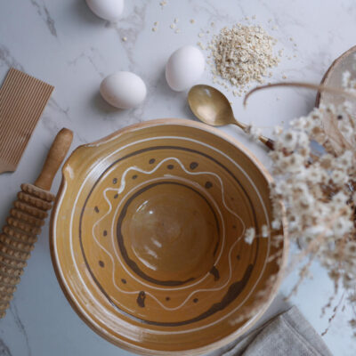 Top-down view of a decorative ceramic mixing bowl with concentric brown patterns, surrounded by eggs, oats, a spoon, and a honey-dipped roller on a marble surface.