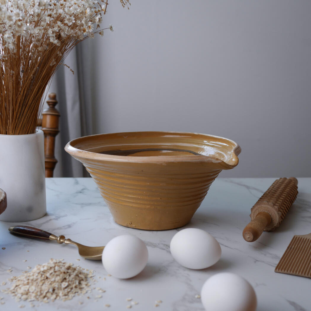 Tan glazed mixing bowl on a marble counter with eggs, oats, a whisk, and a rolling pin ready for baking