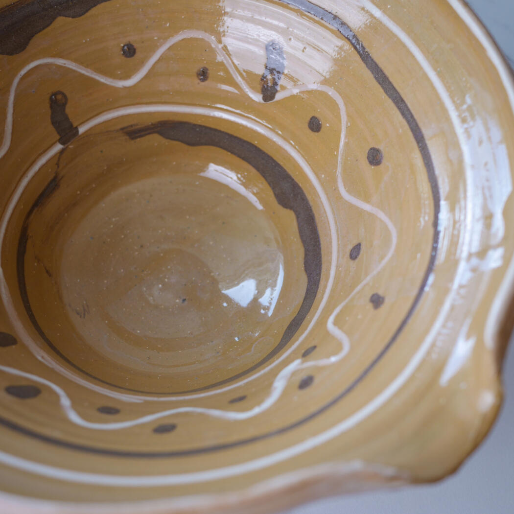 Close-up of a brown-yellow glazed ceramic bowl interior with white wavy lines and dark dots in a concentric pattern.