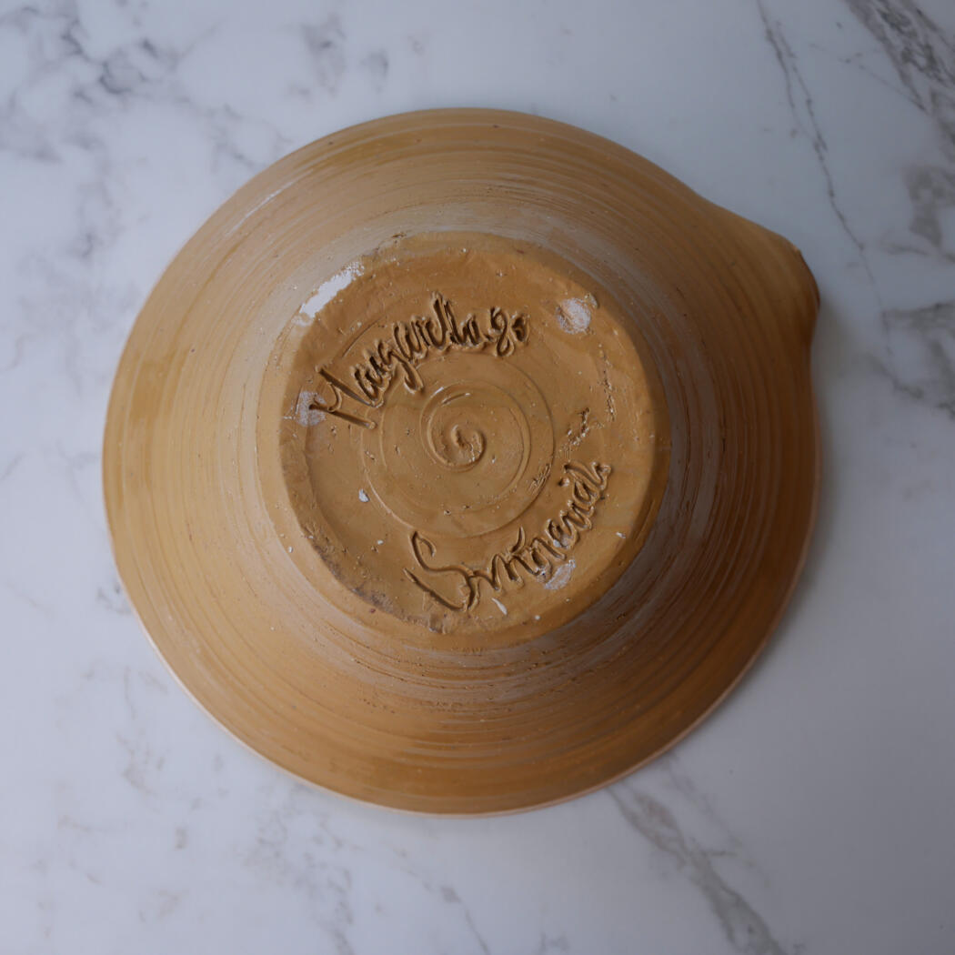 Bottom view of a brown ceramic plate showing a raised, cursive maker's signature embossed in the center of the base.