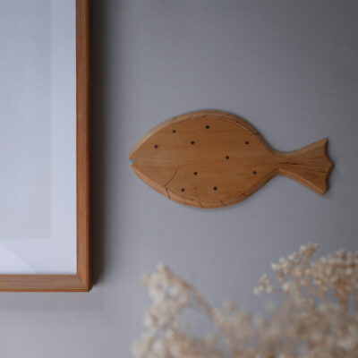 Wooden fish wall decor on a gray wall beside a framed picture frame, with dried flowers in the foreground.