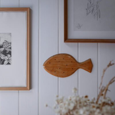 Wooden fish-shaped wall decor mounted between framed prints on a white paneled wall.