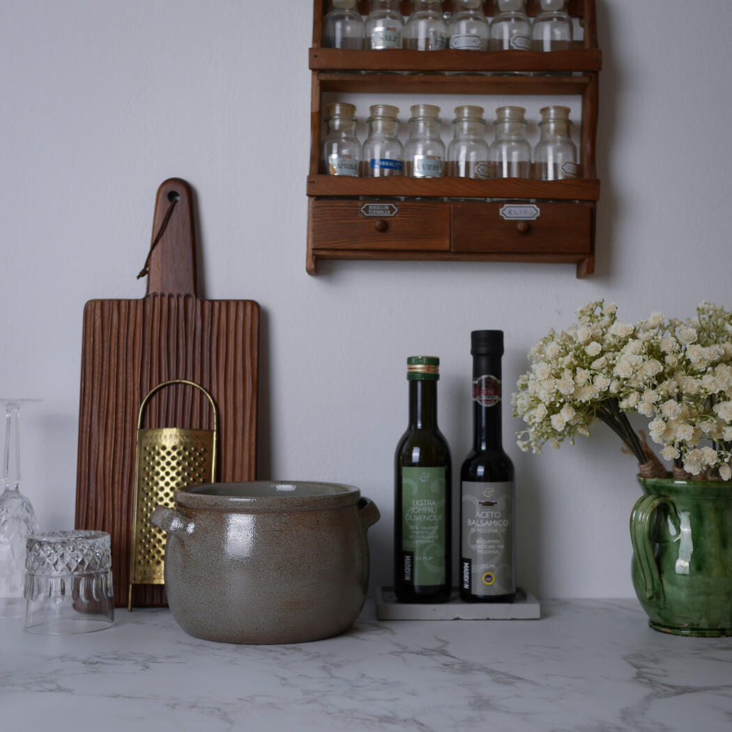 Ceramic pot on a marble countertop beside a wooden cutting board, a spice rack shelf, two olive oil bottles, and a green vase with white flowers.