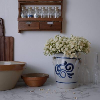 White bouquet in a blue-and-white ceramic pitcher on a marble counter, with a wooden spice rack and glass bottles in the background.