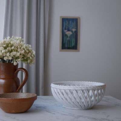 White woven basket on a marble table beside a brown ceramic pitcher with cream flowers, and a brown bowl; gray curtain and wall art in background.