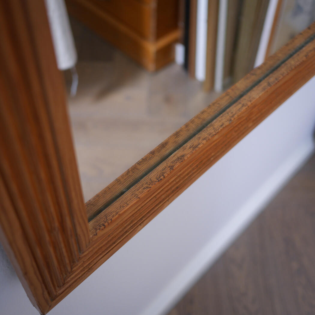 Close-up of a wooden-framed mirror mounted on a wall, reflecting part of a room with shelves behind it.